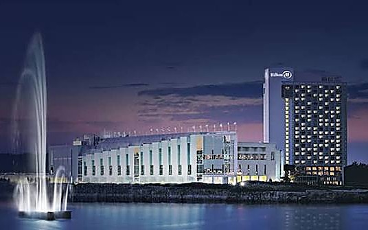Torre de hotel iluminada junto a un edificio bajo de vidrio sobre el agua, al anochecer, con un chorro de agua en primer plano.